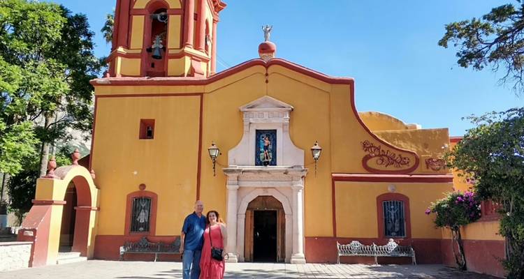 Two visitors stand in front of a mustard-yellow colonial church with a small bell tower under clear blue skies.