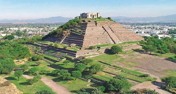 Aerial view of a stepped pyramid covered in greenery overlooking surrounding plains and distant mountains.