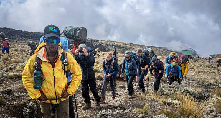 Una fila de excursionistas con mochilas pesadas asciende por las laderas alpinas áridas del Monte Kilimanjaro bajo nubes arremolinadas.