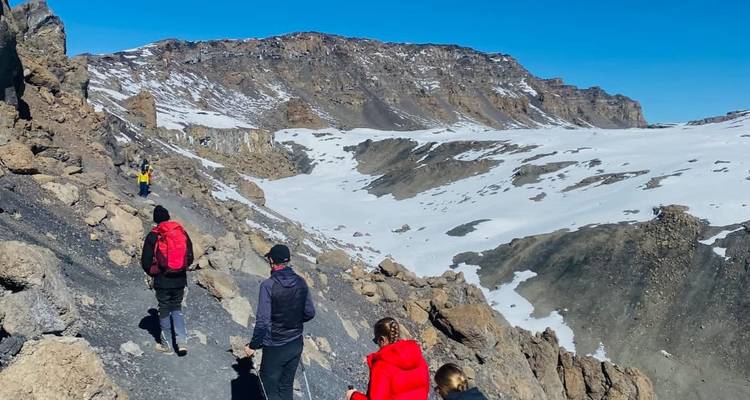 Los montañistas recorren una cresta rocosa con vistas al cráter nevado del Kilimanjaro bajo un sol brillante.