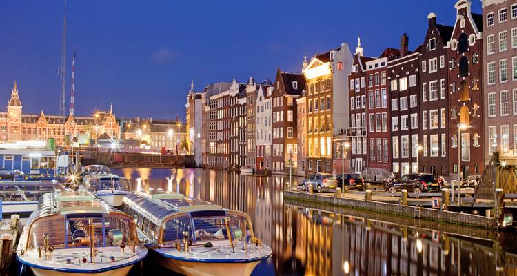 Casas de canal y tejados a dos aguas de Ámsterdam bellamente iluminadas por la noche con barcos turísticos atracados en primer plano.