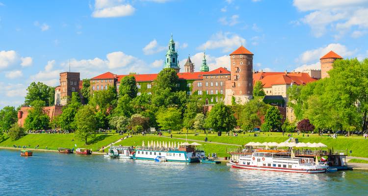 El Castillo de Wawel con vista al río Vístula con barcos turísticos amarrados a lo largo del malecón verde.