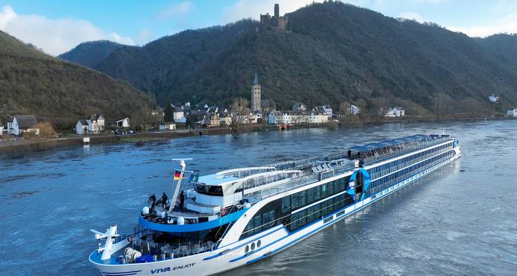 Barco de crucero fluvial moderno y largo deslizándose por un río ancho con ruinas en la cima de una colina y pueblo al fondo.