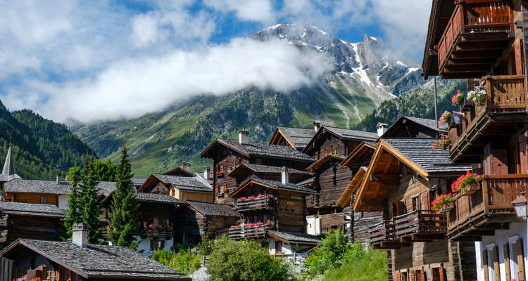 Charmant village alpin de chalets en bois ornés de fleurs sous des pentes verdoyantes et des pics enneigés.