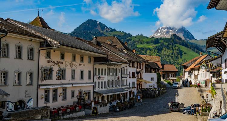 Rue principale historique de Gruyères bordée de restaurants et décor alpin de collines verdoyantes et de sommet enneigé.