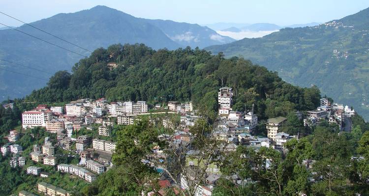 Blick von einem Hügel über dichten Wald zu den zusammengedrängten Gebäuden von Gangtok vor dem Hintergrund dunstig blauer Berge.