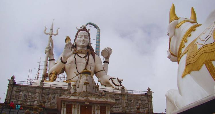 Hoch aufragende weiße Statue von Lord Shiva mit goldenem Stier Nandi dominiert einen Tempelkomplex auf einem Hügel unter bewölktem Himmel.