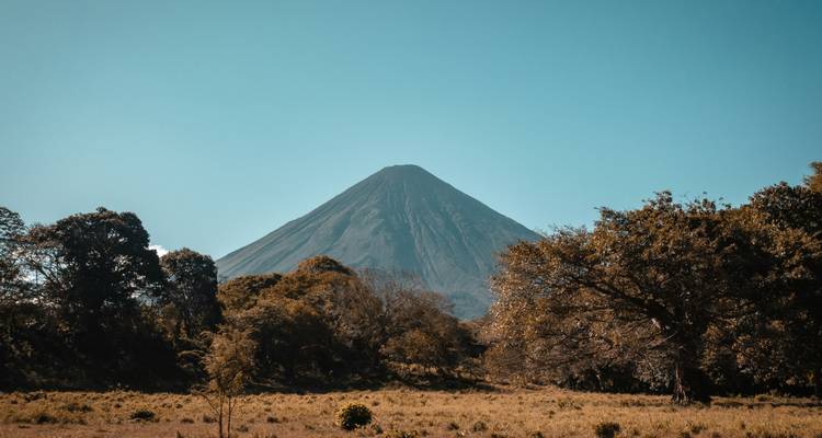 Symmetrical cone volcano rises above wooded plains under a clear blue sky.