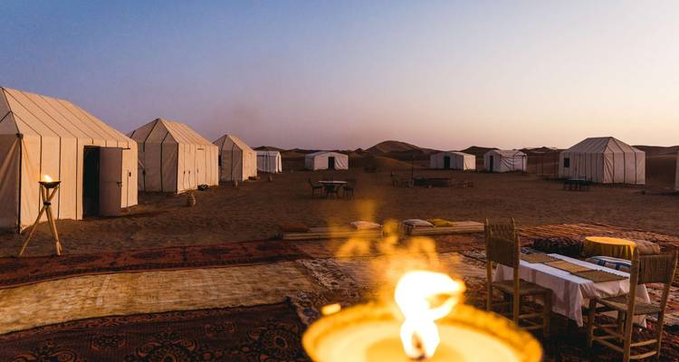 Campamento de glamping en el desierto con tiendas de lona blanca y alfombras iluminadas con velas bajo cielos al atardecer.