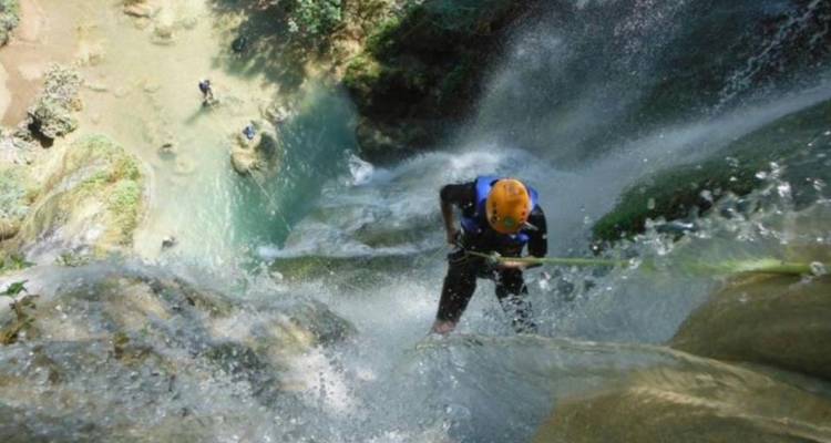 Person seilt sich einen mächtigen Wasserfall hinab, während Wasser überall umherspritzt in einer üppigen Schluchtlandschaft