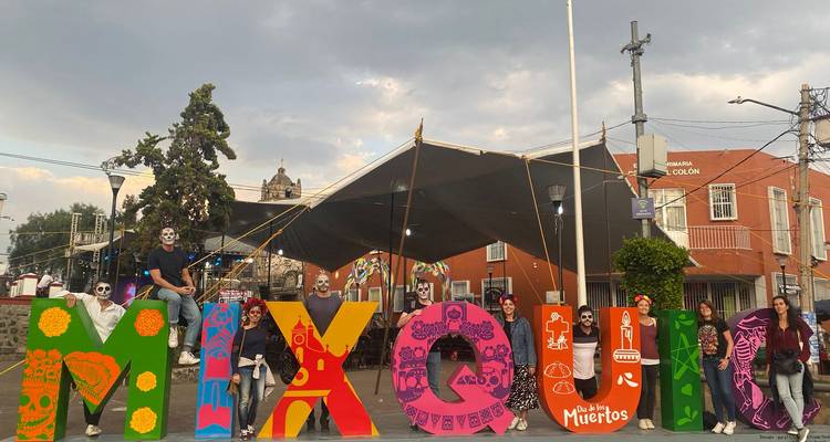 Mensen met Dag van de Doden gezichtsverf poseren naast een kleurrijk 'MIXQUIC' bord op het plein