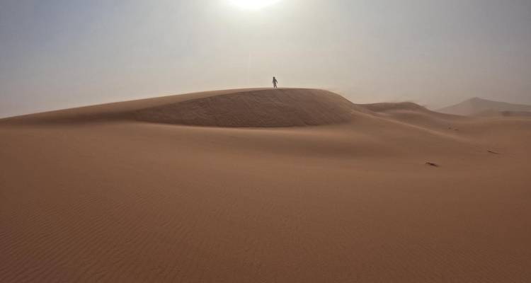 Een eenzame figuur staat bovenop een uitgestrekte door de zon geblakerde zandduinen onder een wazig middagzon in de Sahara.