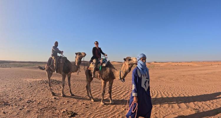 Gids leidt twee ruiters op kamelen door zonovergoten woestijnzand met verre duinen aan de horizon.