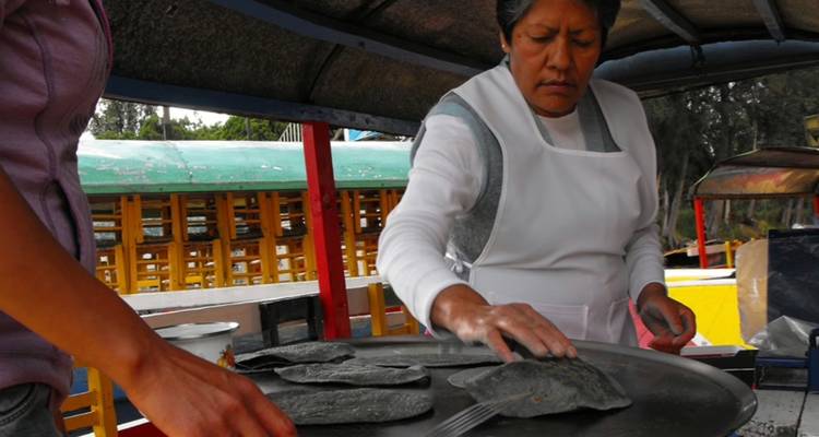 Mujer local con delantal prepara tortillas de maíz azul en una plancha caliente a bordo de una embarcación de canal.