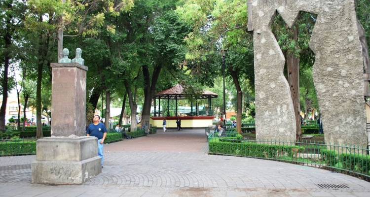 Parque urbano sombreado con estatuas, árboles maduros y lugareños relajándose en bancos alrededor de un quiosco central.