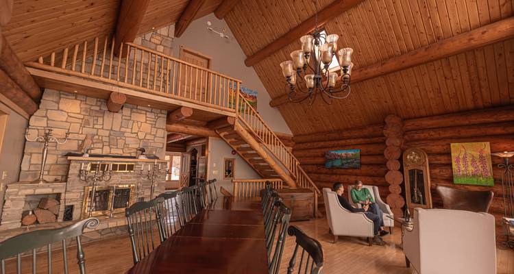 Intérieur chaleureux de chalet en rondins avec cheminée en pierre, longue table à manger et couple conversant dans des fauteuils