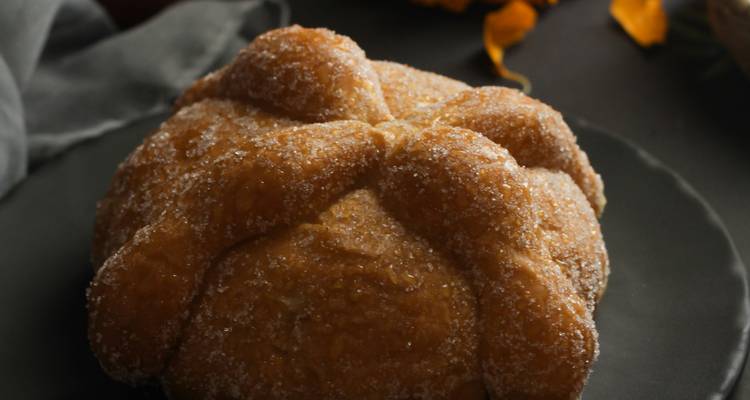 Close-up van een met suiker bestoven Pan de Muerto broodje op een donker bordje met goudsbloemblaadjes ernaast.