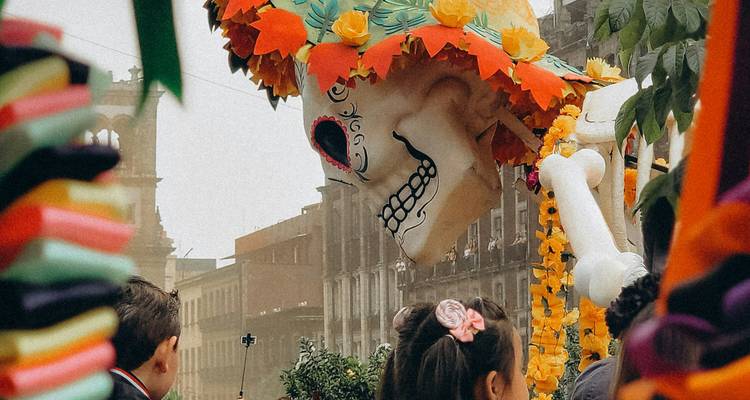 Groot papier-mâché skeletfiguur met een bloemen-sombrero dat door een feestelijke straatparade beweegt.