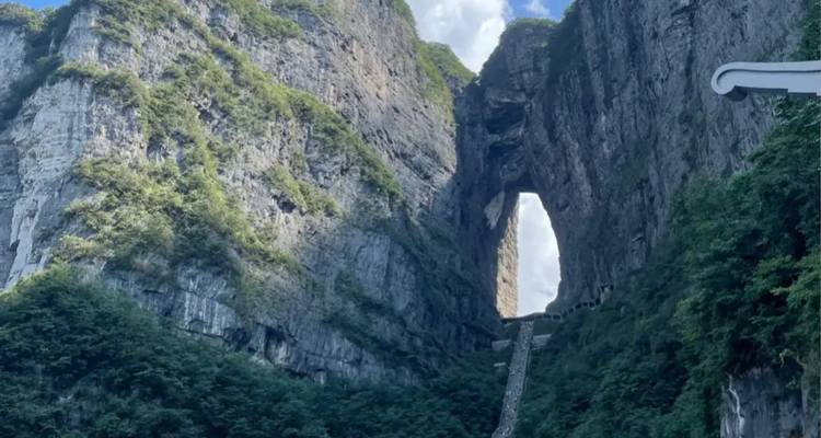 Sheer limestone cliffs rise on both sides of a massive natural archway with a long stairway leading up to it under a blue sky.