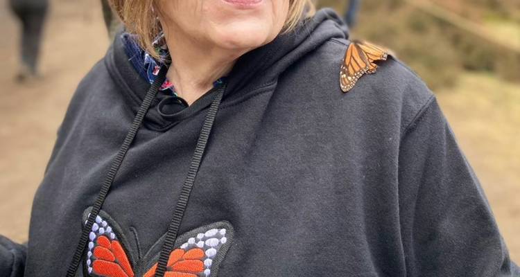 Una mujer que lleva una sudadera con capucha decorada con motivos de mariposas observa mientras una monarca viva descansa en su hombro.