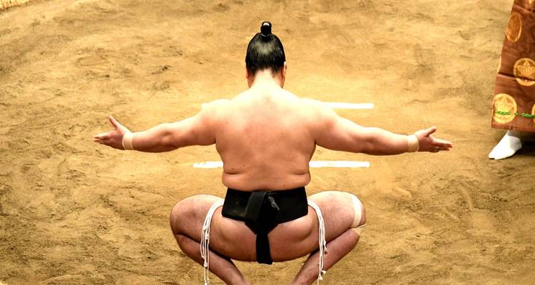 Sumo wrestler performing ceremonial squat in ring on earthen arena floor.