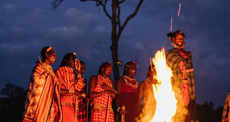 Maasai people in traditional attire singing and dancing around a blazing campfire at dusk.