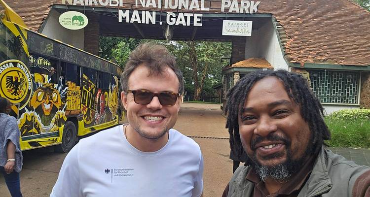 Two travellers smiling in front of Nairobi National Park main gate entrance.