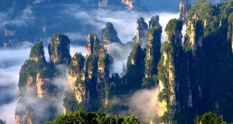 Dramatic pillar-like sandstone peaks rise above mist-filled ravines in a lush green mountainous landscape.