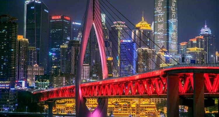 A striking red bridge spans a river with the illuminated skyscrapers of Chongqing’s skyline behind at night.