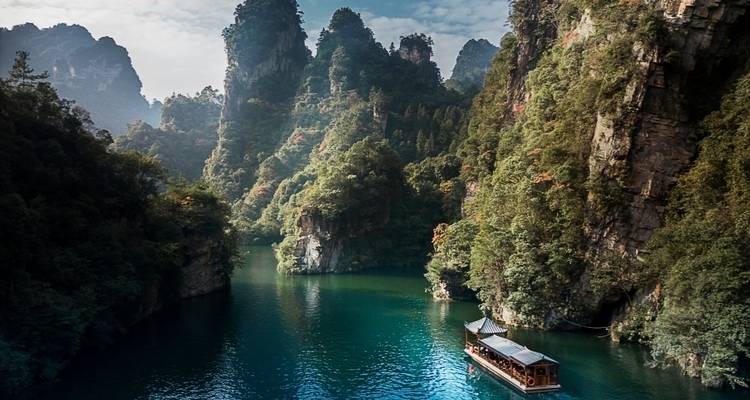 A wooden sightseeing boat glides along emerald water between steep forested karst peaks.