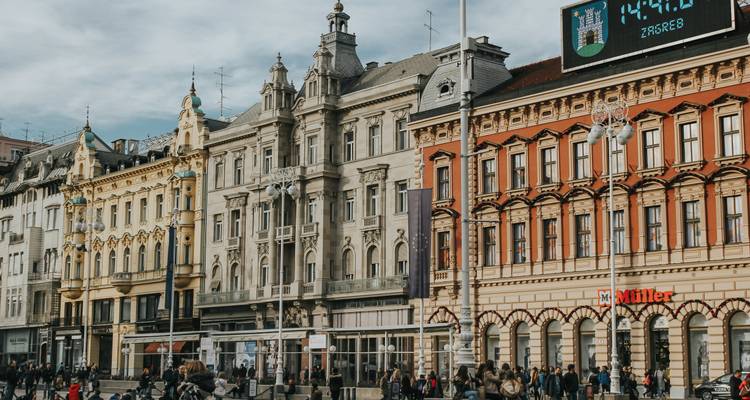 Plaza urbana concurrida en Zagreb bordeada de edificios históricos ornamentados y un reloj digital que muestra el nombre de la ciudad.