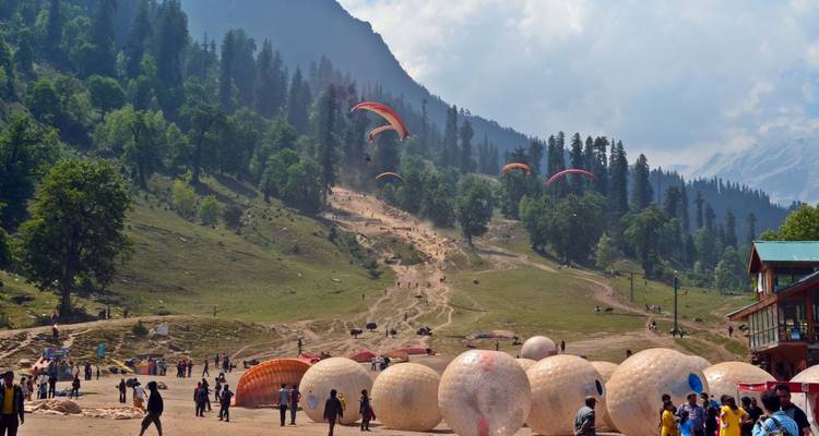 Belebte Bergwiese mit bunten Zorbing-Bällen und mehreren Paragleitern, die über einen von Kiefern gesäumten Berghang nahe dem Himalaya schweben.