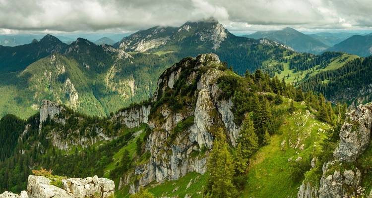 Weitläufiges Panorama zerklüfteter grüner Berge unter stimmungsvollen Wolken mit gestaffelten Gebirgszügen dahinter.