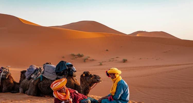 Dos hombres bereber con turbantes coloridos se sientan junto a camellos contra dunas de un naranja ondulante al amanecer.