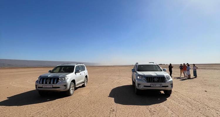 Zwei 4x4 SUVs geparkt auf einem weiten, rissigen Wüstenseebett, während Reisende in der Ferne unter klarem blauen Himmel stehen.