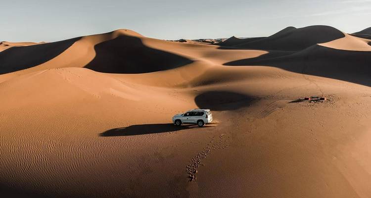 Drohnenperspektive eines einsamen SUV, der bei Sonnenaufgang die hoch aufragenden Sanddünen in der weiten Erg Chigaga Wüste durchquert.