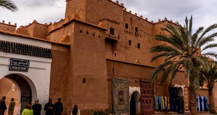 Historische Adobe-Kasbah mit Palmen und bunten Teppichen, die draußen ausgestellt sind, Besucher betreten den Eingang.