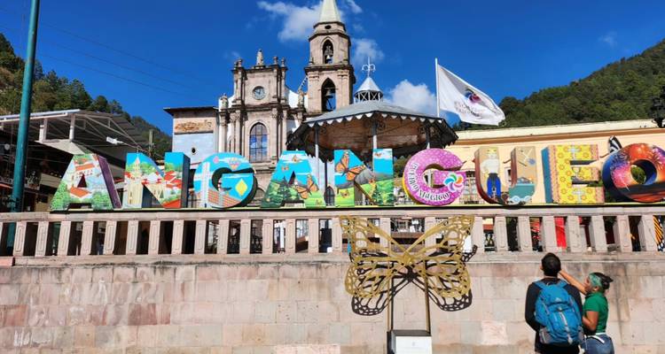 Letrero colorido de ANGANGUEO con iglesia colonial y cielo azul mientras dos turistas lo admiran