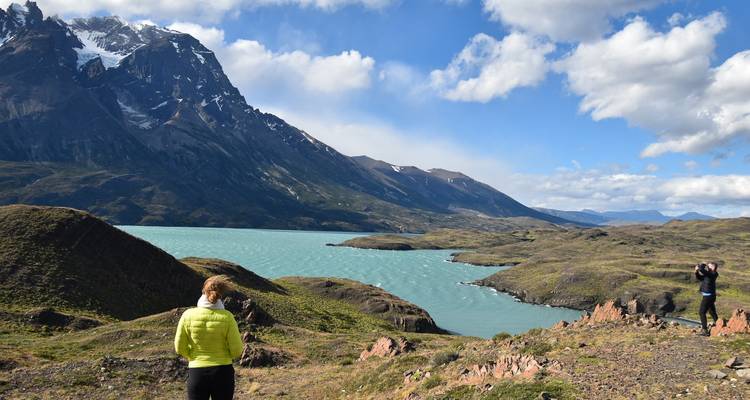 Dos viajeros admiran un lago turquesa enmarcado por picos andinos escarpados y un cielo parcialmente nublado.
