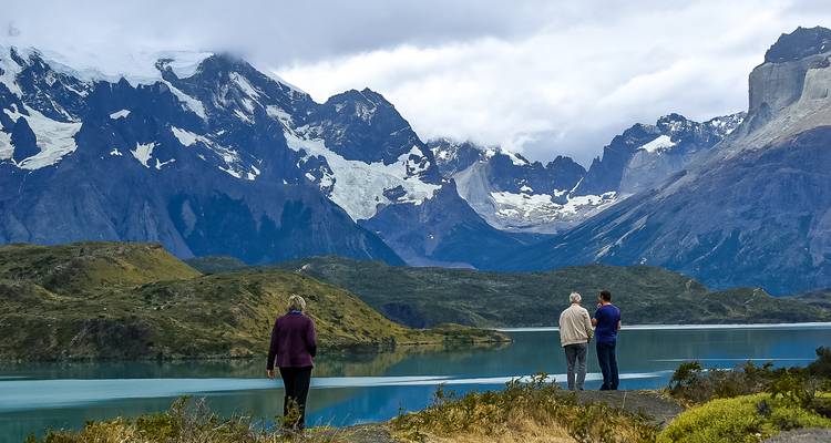 Tres visitantes se paran en la orilla de un lago patagónico reflectante dominado por montañas glaciares.