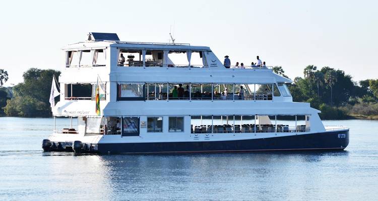 Zweistöckiges weißes Kreuzfahrtschiff gleitet auf einem ruhigen Fluss mit Passagieren auf dem Oberdeck