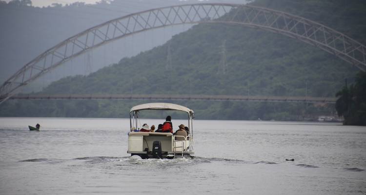 Kleines Motorboot mit Passagieren auf einem breiten Fluss mit einer eleganten Bogenbrücke und grünen Hügeln im Hintergrund.