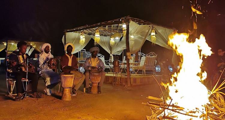 Musiker in traditioneller Kleidung trommeln und singen neben einem lodernden Lagerfeuer in einem Wüstenlager bei Nacht.