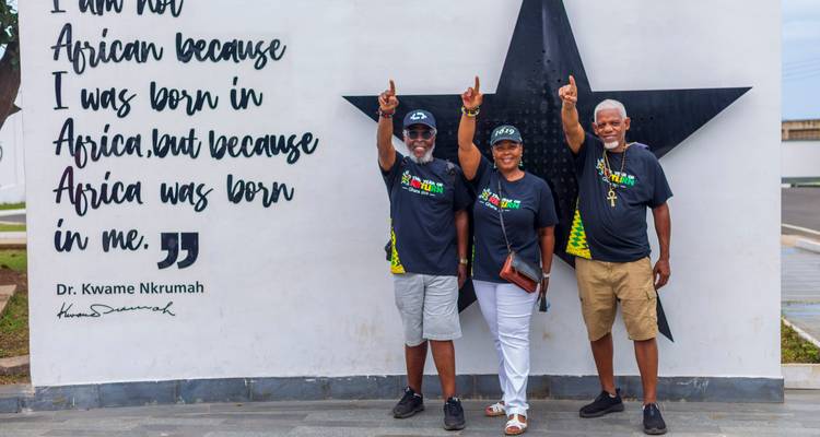 Trois voyageurs souriants posent devant une fresque murale géante représentant une étoile noire citant le Dr Kwame Nkrumah.