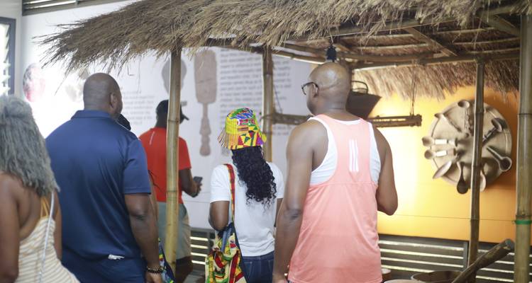 Un groupe de touristes écoute un guide à l'intérieur d'une hutte d'exposition au toit de chaume rustique présentant des objets traditionnels.
