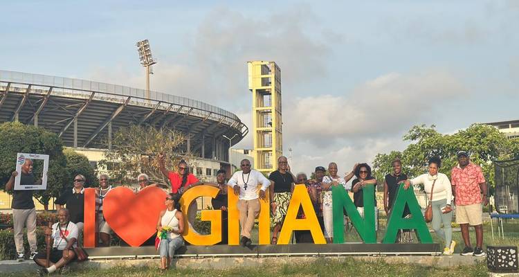 Un grand groupe de touristes pose avec des lettres colorées « I ❤️ GHANA » devant un stade sportif.