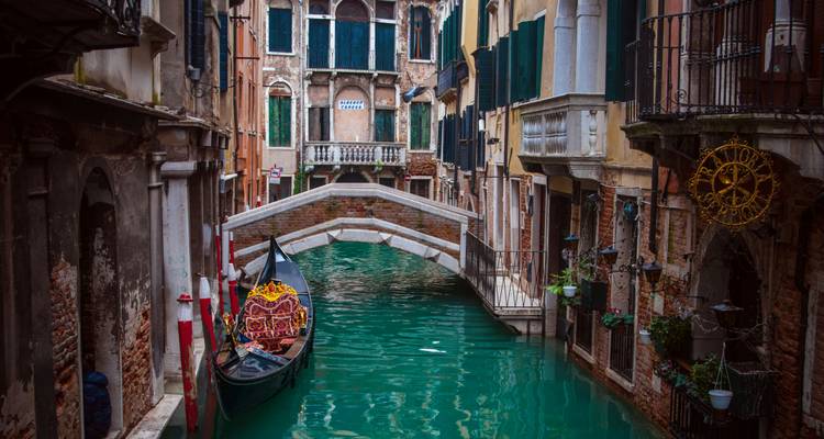 Canal estrecho y pintoresco en Venecia con una góndola decorada amarrada bajo un pequeño puente de piedra entre edificios históricos.