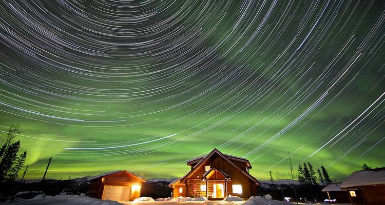 Cabañas de troncos bajo senderos de estrellas giratorias y vívidas auroras boreales verdes en la noche de invierno de Yukón.