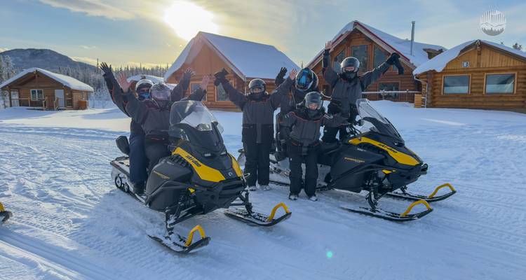 Grupo de viajeros posando en motos de nieve frente a cabañas de madera en un día soleado de invierno.