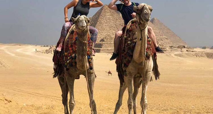 Dos turistas montando camellos frente a las Pirámides de Giza en el desierto.
